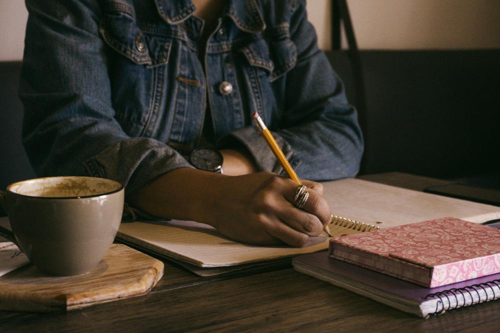 Foto: Vrouw schrijft in een schrift, met agenda en kopje koffie ernaast.