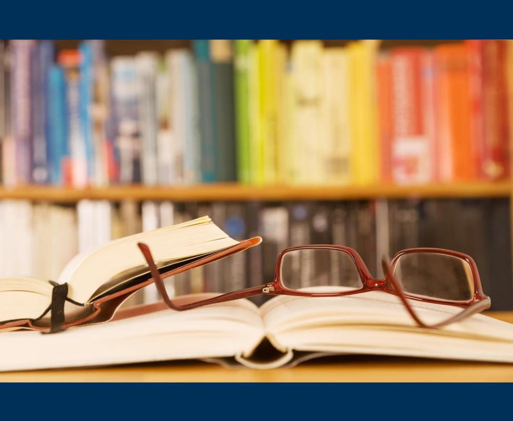 Op de voorgrond liggen twee opengeslagen boeken met daarop een leesbril. Op de achtergrond staat uit focus een op kleur gerangschikte boekenkast.