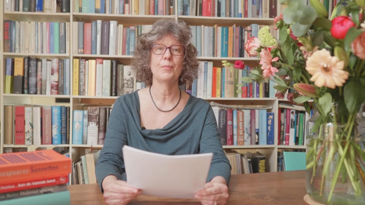 Vertaler Jacqueline Hulst zit aan tafel met naast haar de vijf genomineerde vertalingen en kijkt in de camera. Achter haar staat een boekenkast gevuld met boeken.