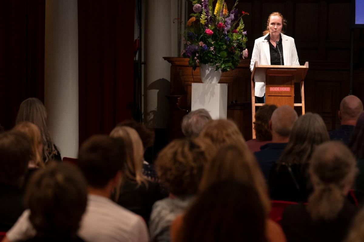 Foto van Marije de Bie achter het spreekgestoelte, gezien vanuit het publiek. 